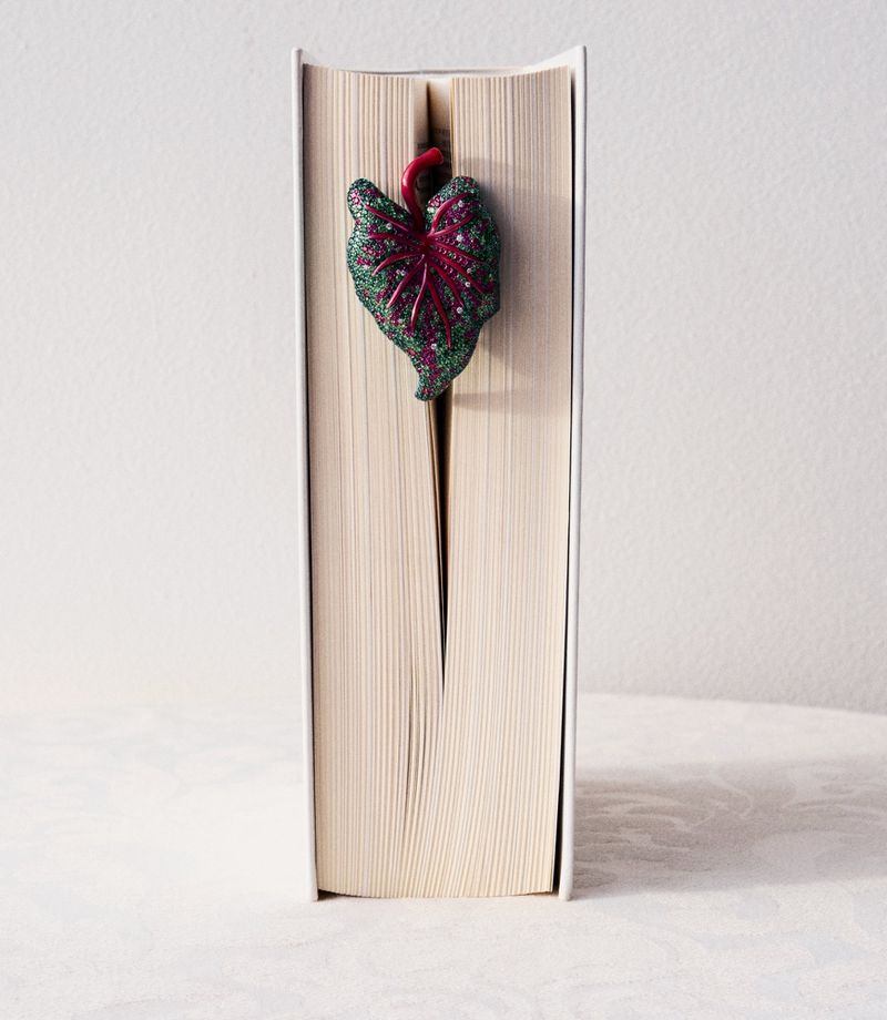 Book with white cover and a decorative leaf bookmark with dark green and red tones standing upright on a neutral surface.