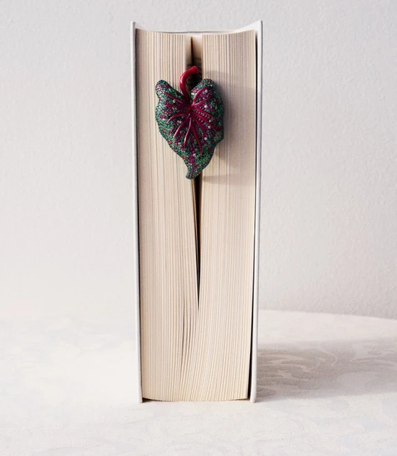Book with white cover and a decorative leaf bookmark with dark green and red tones standing upright on a neutral surface.