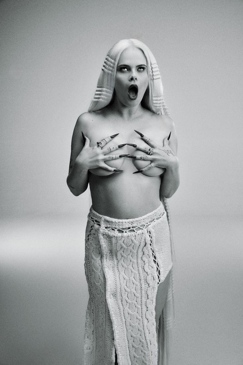 Striking black-and-white portrait of a woman with long blonde hair, dark eye makeup, and long black nails, posing in a knitted skirt.