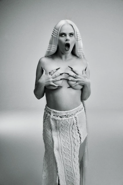 Striking black-and-white portrait of a woman with long blonde hair, dark eye makeup, and long black nails, posing in a knitted skirt.
