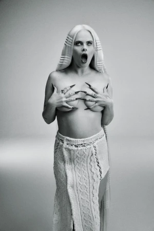 Striking black-and-white portrait of a woman with long blonde hair, dark eye makeup, and long black nails, posing in a knitted skirt.