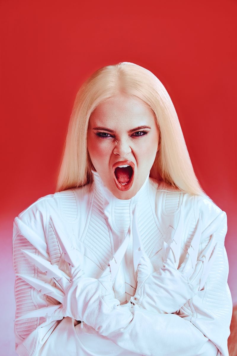 Portrait of a blonde adult female shouting and pulling at a white jacket against a red background in a photo studio with bright lighting.