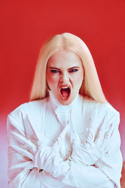 Portrait of a blonde adult female shouting and pulling at a white jacket against a red background in a photo studio with bright lighting.