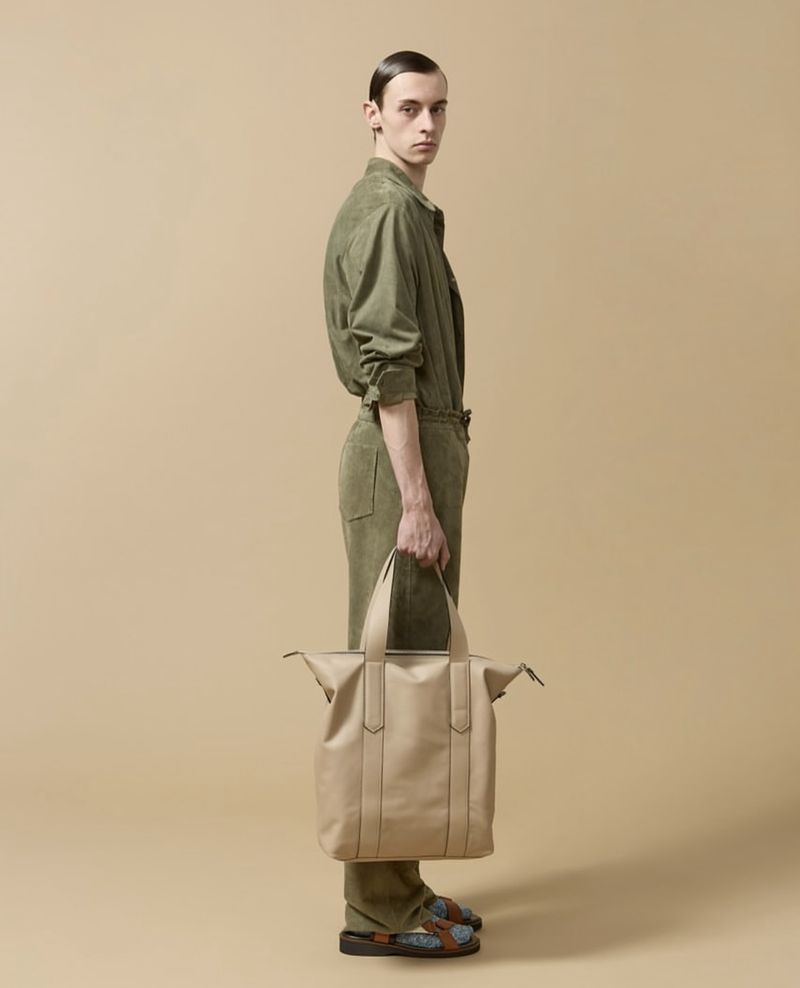 Studio shot: slender adult male poses in an olive green jumpsuit with beige leather bag, standing against a neutral tan backdrop.