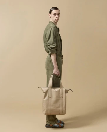 Studio shot: slender adult male poses in an olive green jumpsuit with beige leather bag, standing against a neutral tan backdrop.