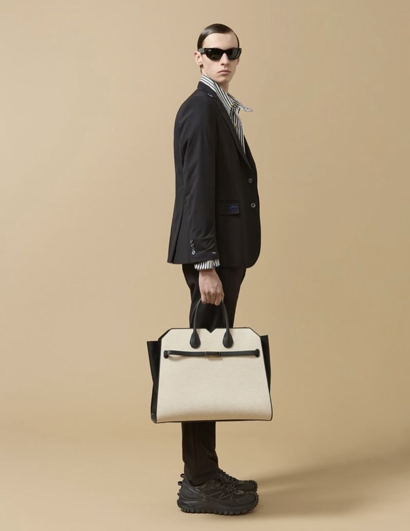A stylish adult male in a suit stands in a studio with a luxury tote bag, black sunglasses, and running shoes against a neutral backdrop.