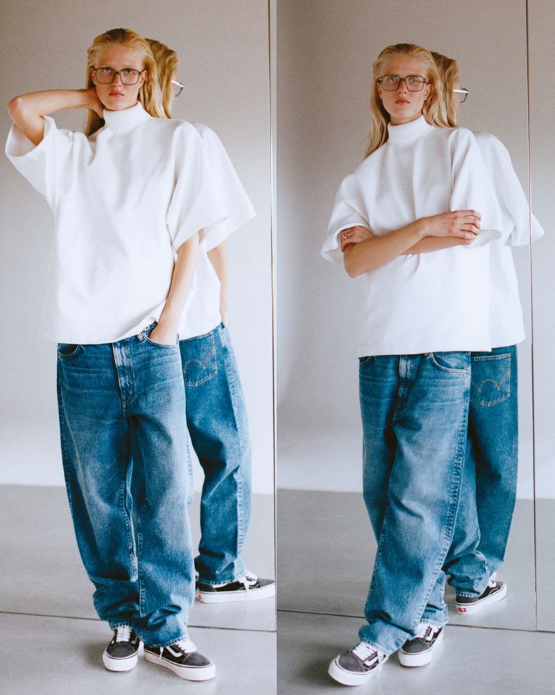 A fair-skinned adult woman in a white top and blue jeans poses in front of a mirror, reflecting her stance, in a studio setting.