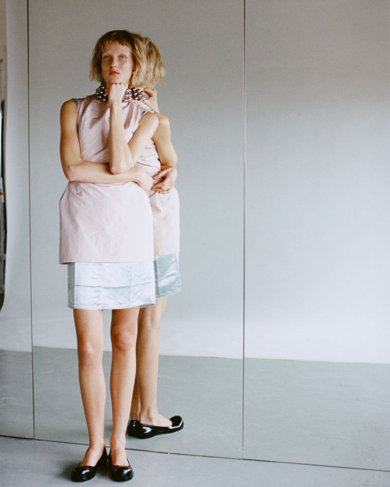 Woman in a pink and blue dress stands near a large mirror with reflection, wearing black flats and a pearl necklace, studio lighting.