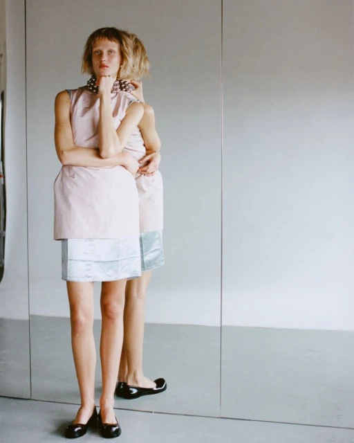 Woman in a pink and blue dress stands near a large mirror with reflection, wearing black flats and a pearl necklace, studio lighting.