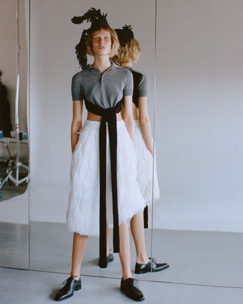 A model poses in an indoor studio with a white background, wearing a grey top, white feathered skirt, and black dress shoes.