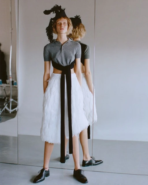 A model poses in an indoor studio with a white background, wearing a grey top, white feathered skirt, and black dress shoes.