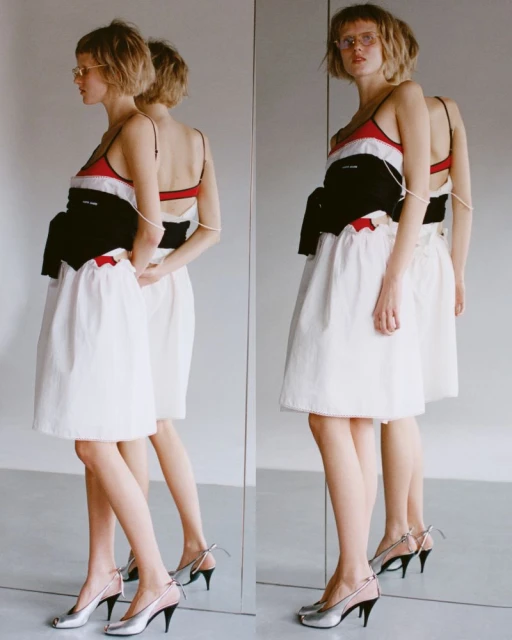 Adult woman posing in a studio next to a mirror. She is wearing a white dress, a black sweater, and silver heels. Editorial fashion shot.