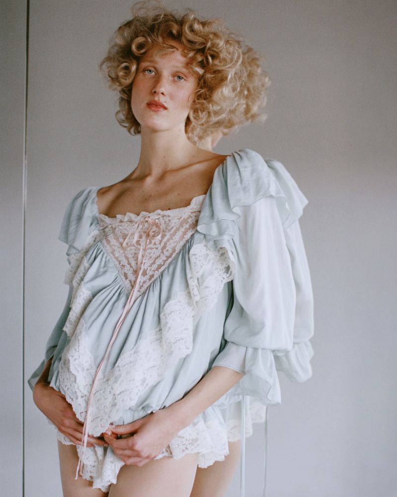 Portrait of a curly-haired woman wearing a blue and white lacy top, posing indoors with hands clasped and a soft, natural light.
