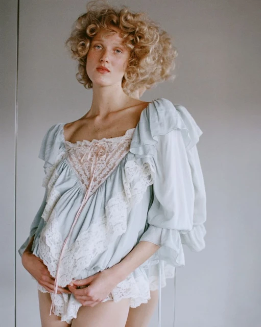 Portrait of a curly-haired woman wearing a blue and white lacy top, posing indoors with hands clasped and a soft, natural light.