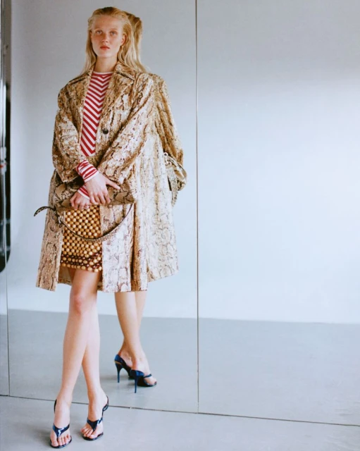 Fashionable woman in patterned trench coat holding handbag, standing in front of mirror, wearing heels. Indoor fashion portrait.
