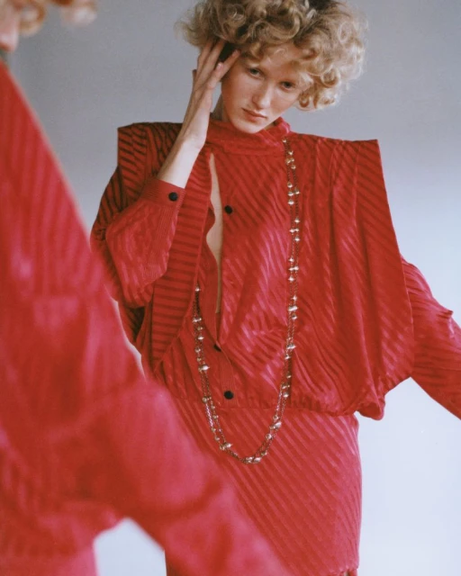 A woman in a red, diagonally striped dress poses in front of a mirror, inside, with her hand near her head and long necklace.