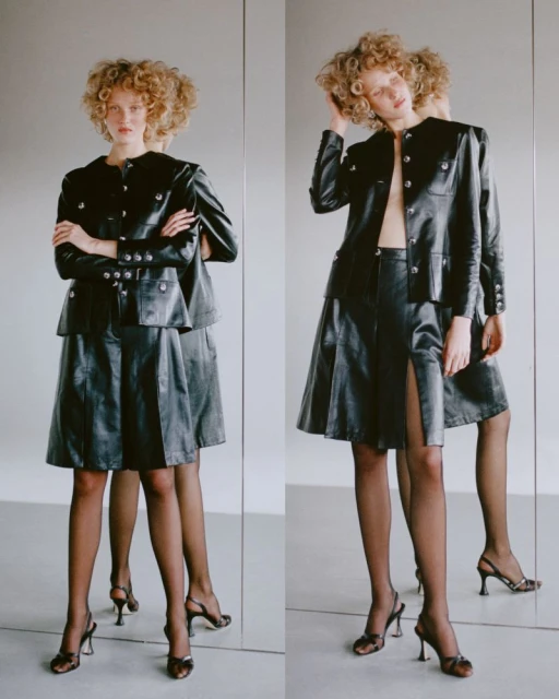Two young women with curly hair pose in black leather skirt suits and heels, standing against a neutral background with a mirror.