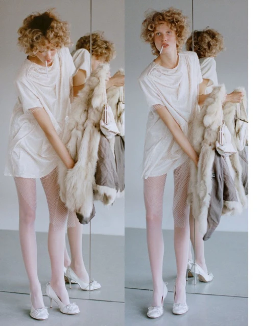 Woman in white dress and fishnet tights posing with a fur coat and heeled shoes next to a mirror in an indoor setting.