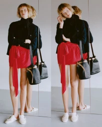 Model wearing a black top and red miniskirt standing next to a mirror reflecting her back, holding a black bucket bag.