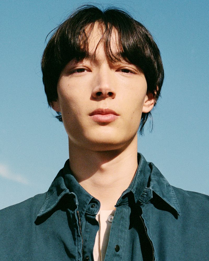 Portrait of a young adult male with dark hair against a clear blue sky, wearing a collared denim shirt.