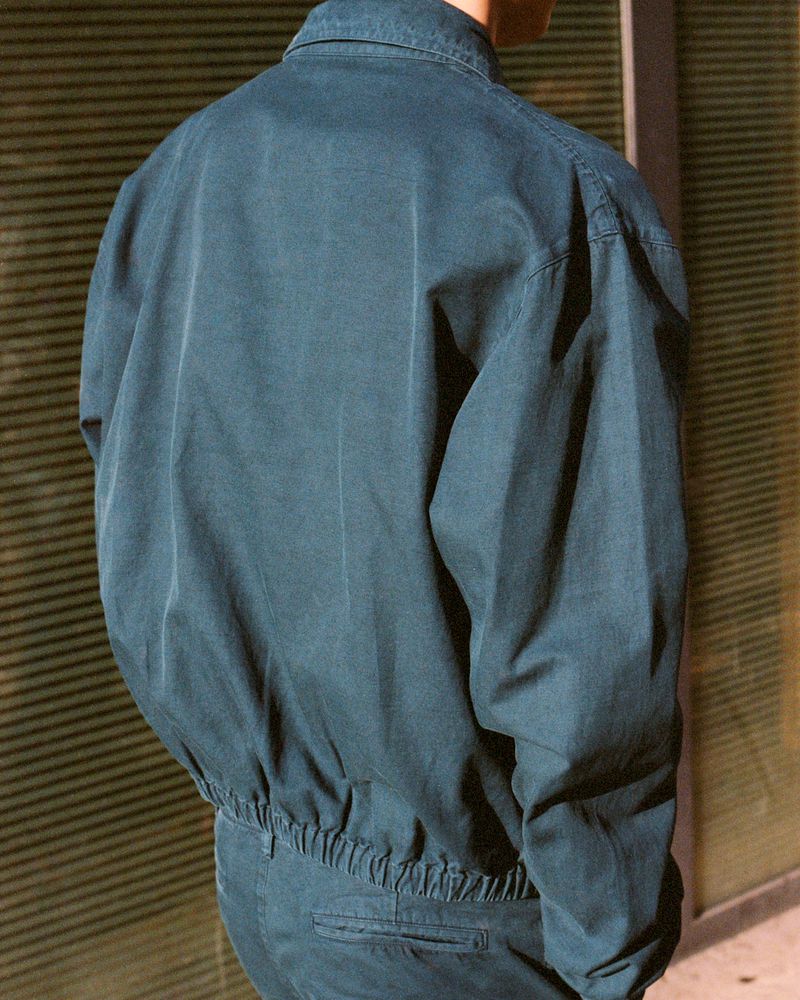 Person wearing a blue collared jacket and matching trousers stands in front of a horizontally striped wall, back to the camera.