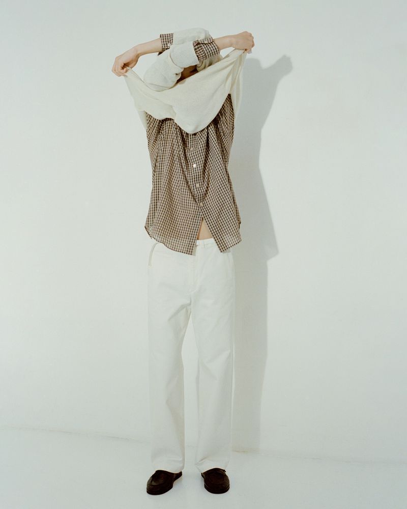 Full-body shot of a human pulling a beige shirt over their head, wearing white pants and brown loafers in a minimalist white studio.