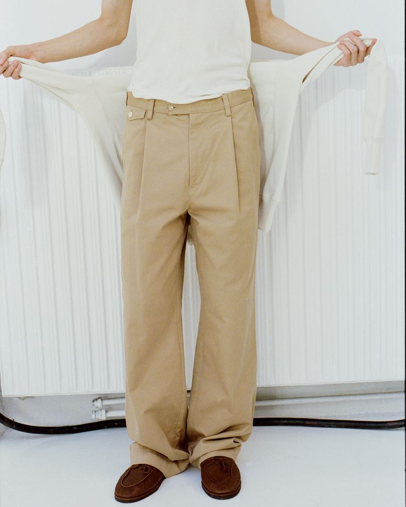 A person stands with wide-leg khaki pants and brown loafers, holding a light-colored sweater in a minimalist indoor setting.