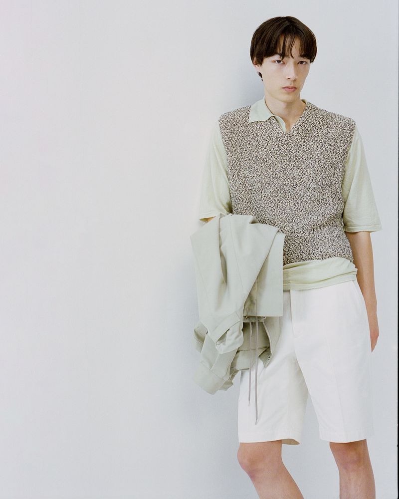 Studio shot of a young adult male wearing a polo shirt, sweater vest, and shorts, holding a jacket, against a solid light-colored background.
