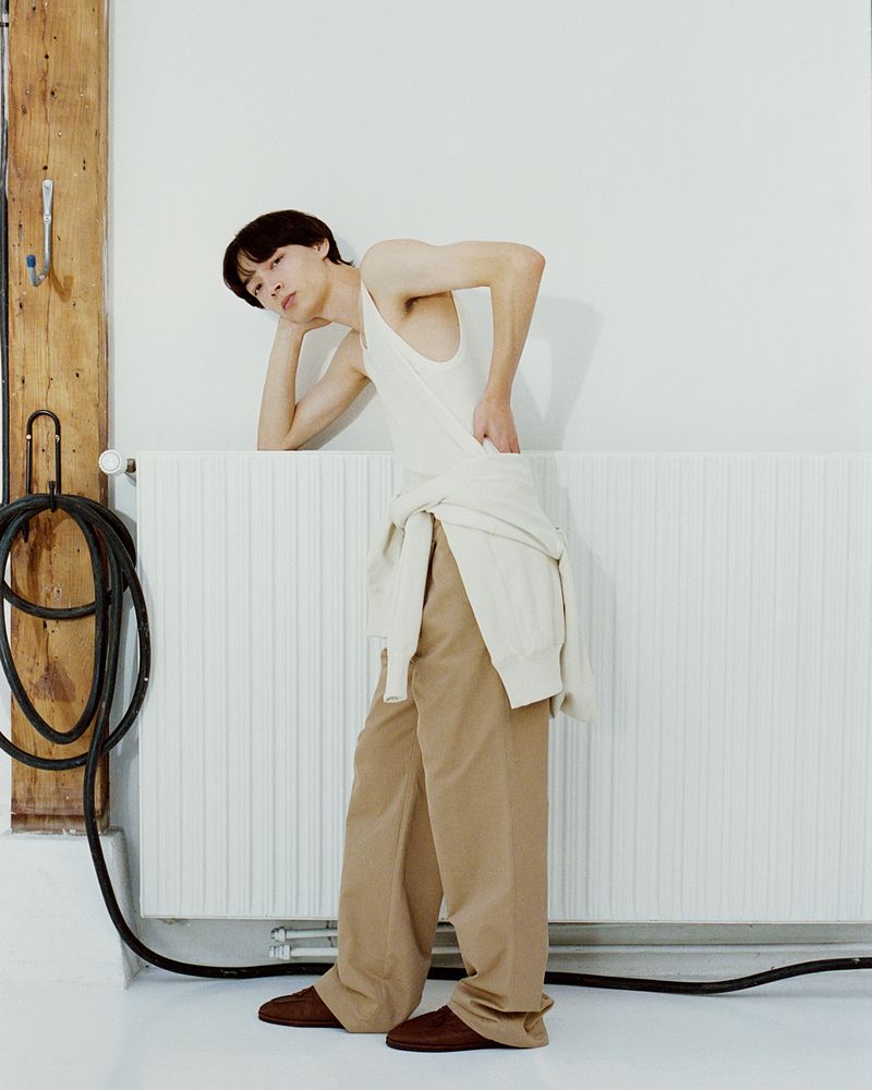 Young adult Asian man in a white tank top and tan pants leaning against a white radiator with a black cord in a minimalist indoor setting.