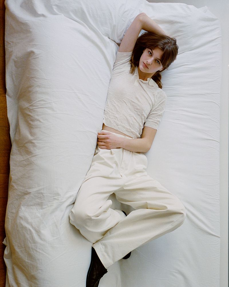 Brunette woman lies on a white bed, wearing a white t-shirt and pants, with dark shoes; taken in an indoor setting.