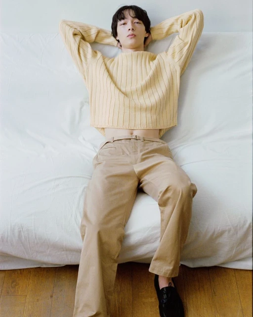 Young male model with dark hair lays on a white bed wearing a yellow ribbed sweater and khaki pants in a fashion shot, looking at camera.