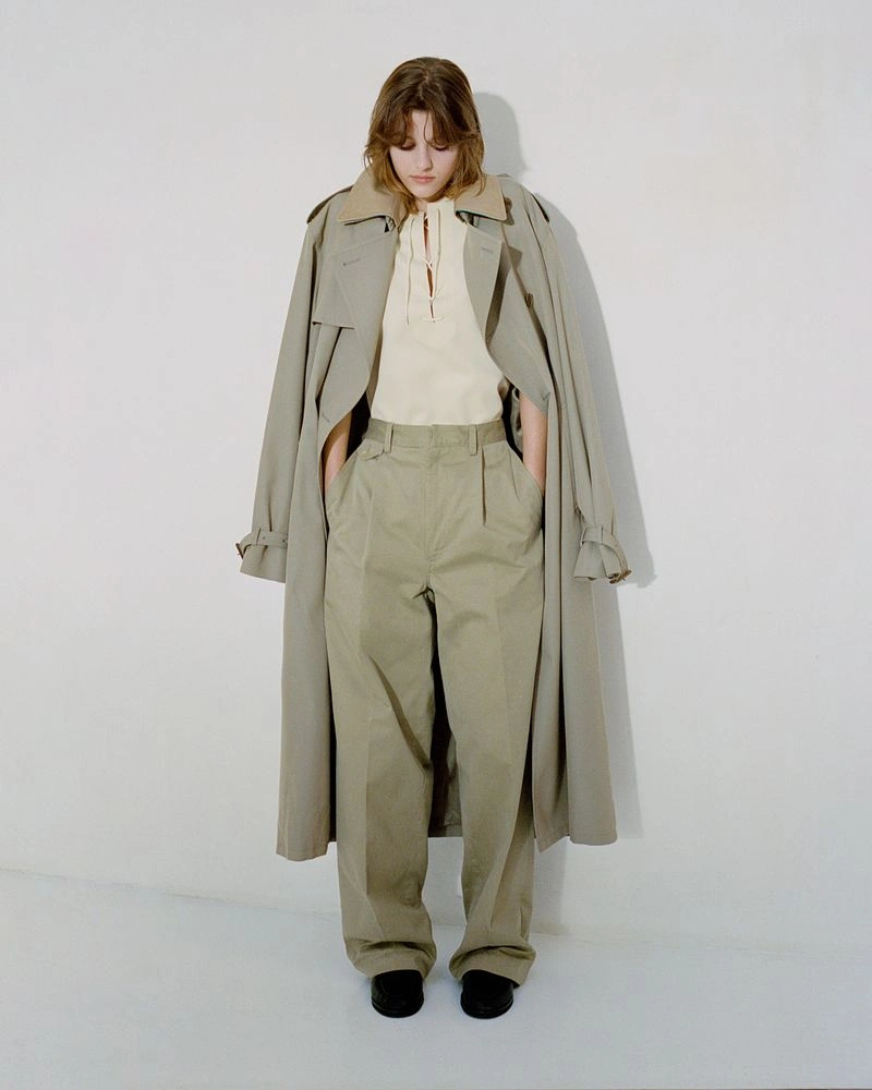 Model wearing a beige trench coat, light yellow top, and pleated trousers against a plain white studio background.