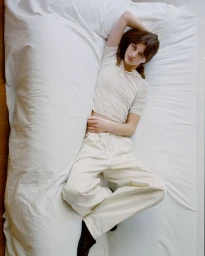 Model lying on a white bed wearing light-colored polo shirt and trousers for Auralee Chino Days campaign.