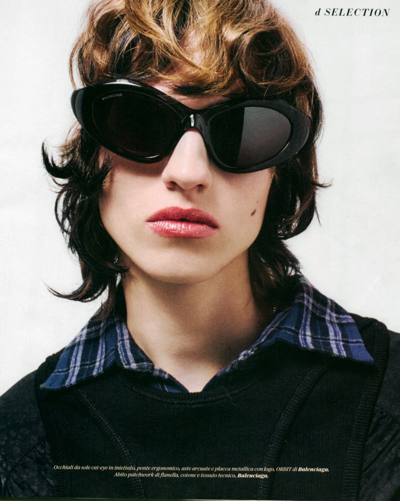 A person with brown curly hair and dark sunglasses wears a black top with a blue plaid collar, shot indoors.