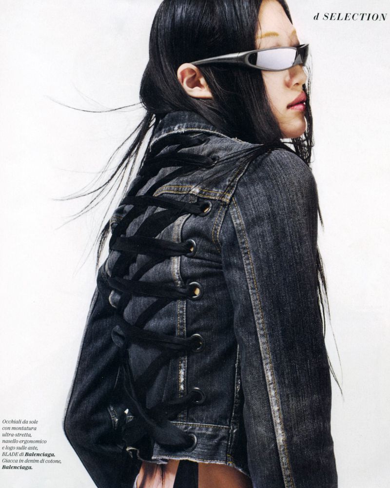Asian woman in sunglasses and denim jacket featuring lace-up back detail, posing against a white background in a fashion shot.