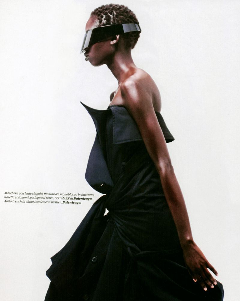 An African American woman models an avant-garde black dress with futuristic visor glasses in a bright, high-fashion studio setting.