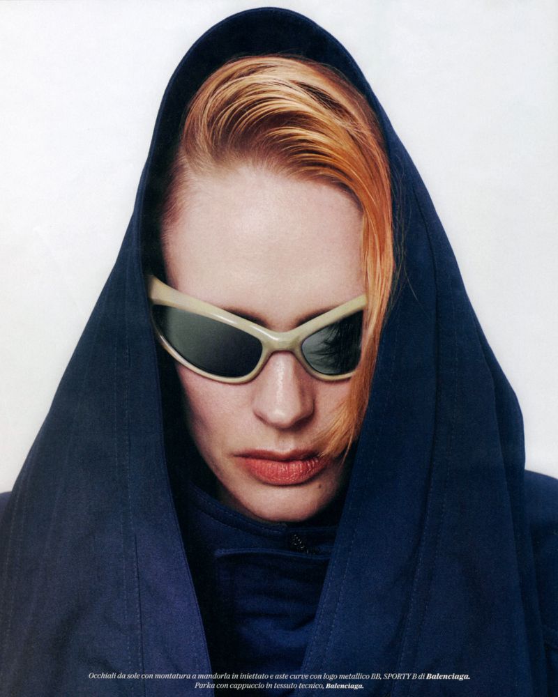 Close-up of a redhead woman wearing futuristic sunglasses and a dark blue hooded top against a white background in a fashion editorial.