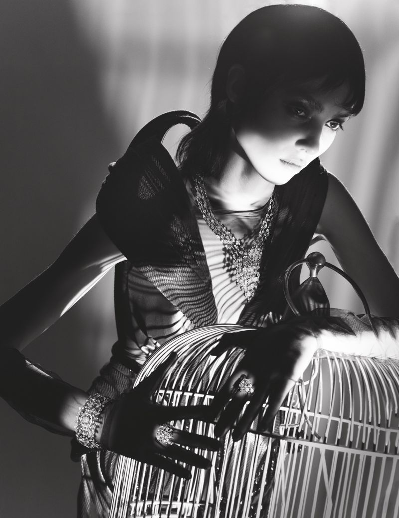 Monochrome portrait of a young woman with short dark hair, wearing jewelry, and holding a birdcage in a dramatic, shadowed light.