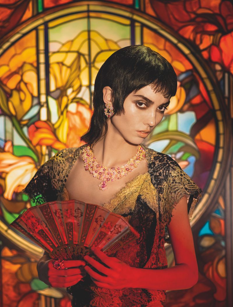 A woman with dark hair and floral jewelry poses with a fan against a colorful stained glass background in a fashion editorial shot.