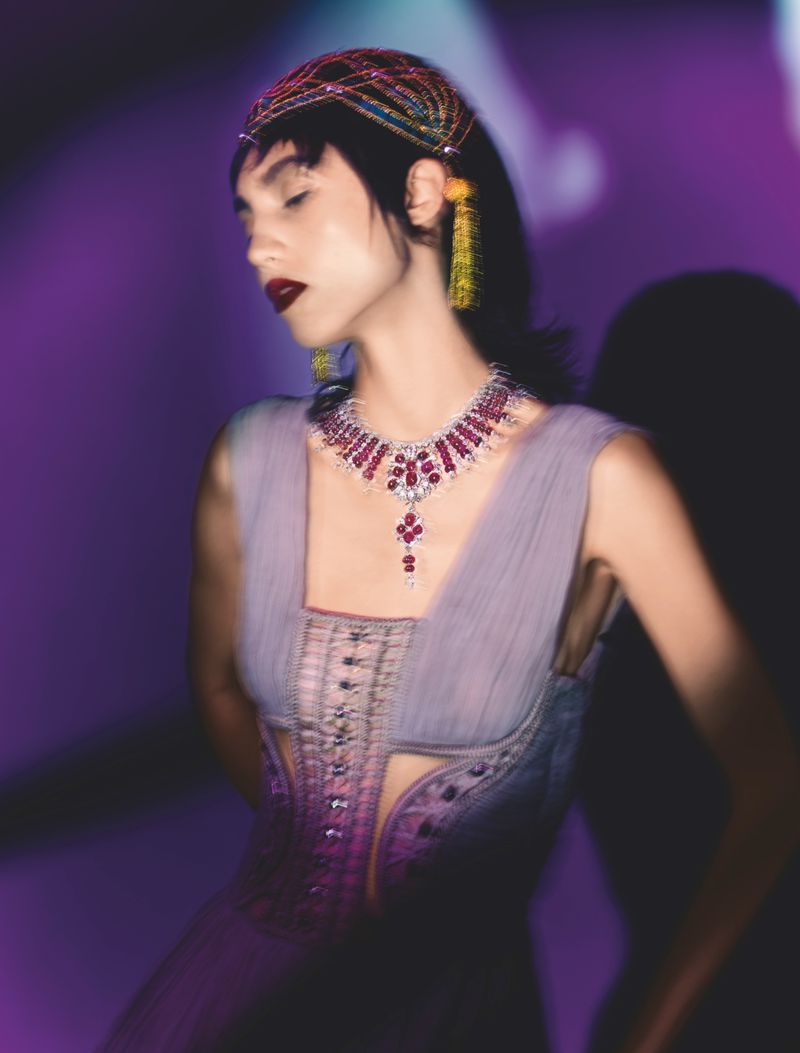 Portrait of a woman with dark hair wearing a jeweled headband, dramatic jewelry, and lavender dress against a purple backdrop.