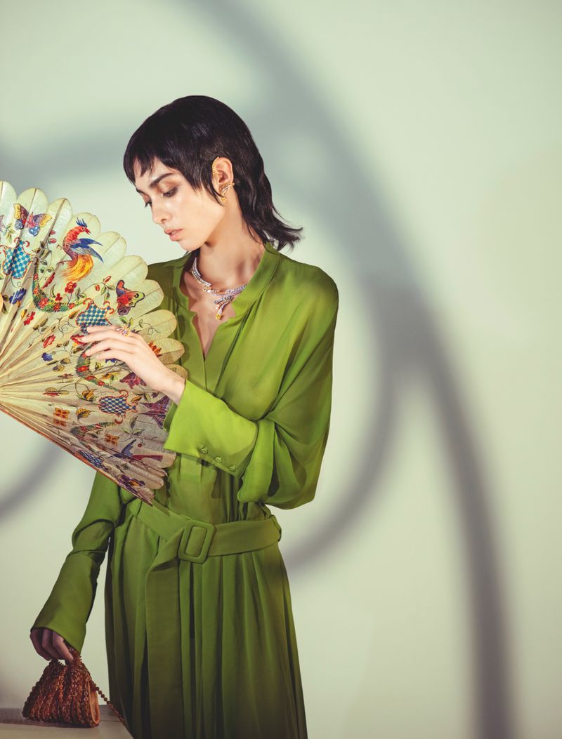 An elegant woman in a green dress holds a decorative fan and a beaded purse in a studio setting, bathed in soft, indirect light.