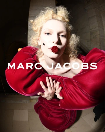 Fashion portrait of a woman with pale skin and curly hair wearing a large red dress and a red, beaded mouth piece against a dark background.