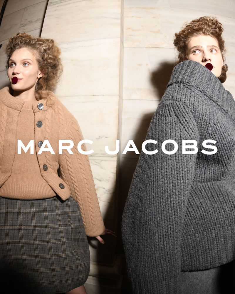 Two fashion models pose indoors wearing Marc Jacobs knitwear; one in tan cable-knit cardigan and plaid skirt, one in chunky grey sweater.