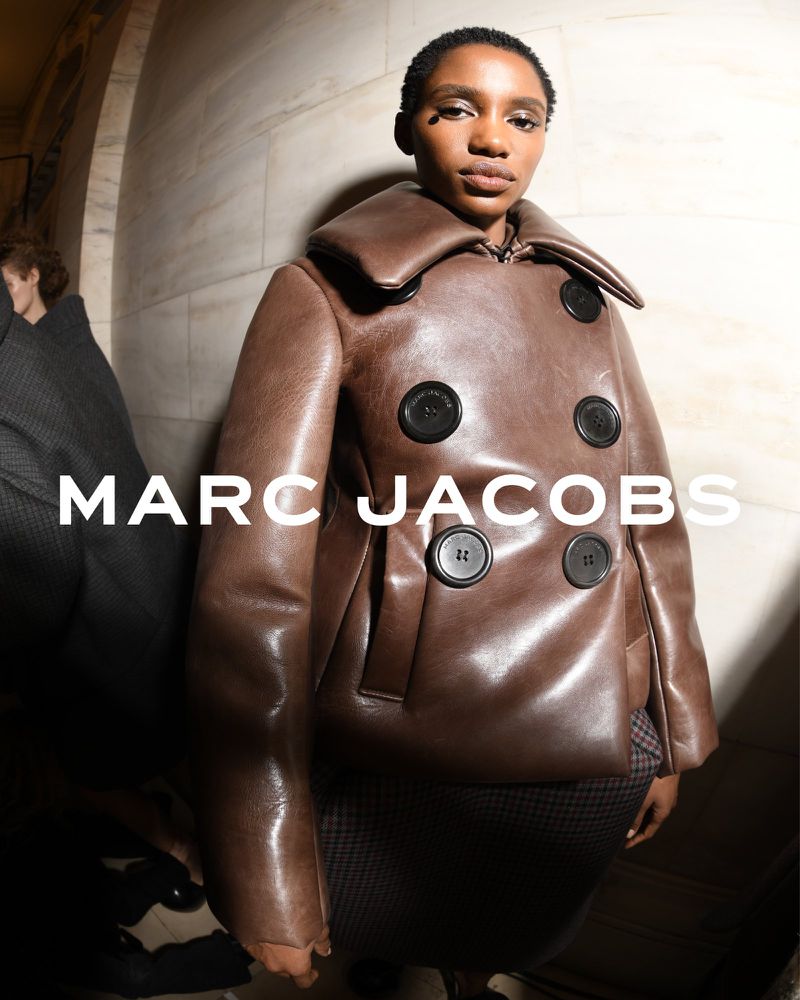 A model poses in a brown leather jacket with large black buttons and a wide collar in a fashion editorial shot with white marble wall.