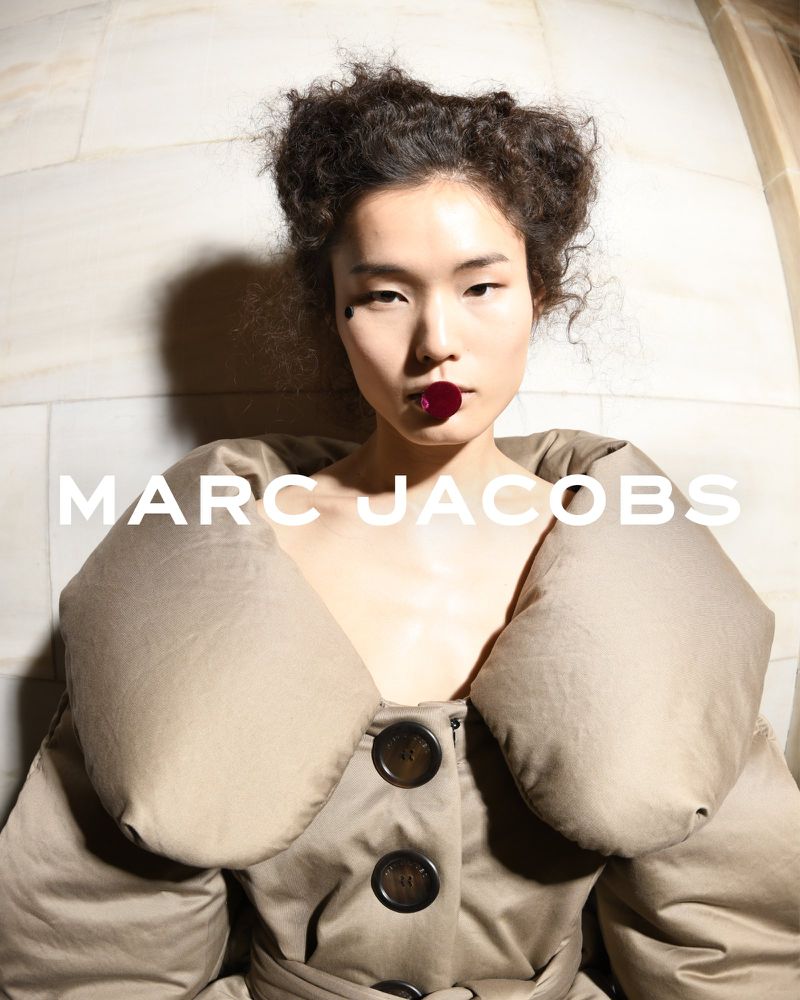 Asian fashion model poses indoors wearing a tan trench coat with a button-up front for Marc Jacobs brand advertising campaign.