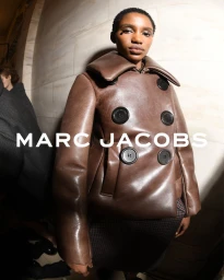 Model wearing a brown leather double-breasted coat with large black buttons against an interior stone wall.