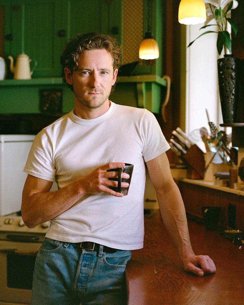 Indoor portrait of a man in a white t-shirt holding a dark mug, leaning against a wooden counter in a warm kitchen setting.