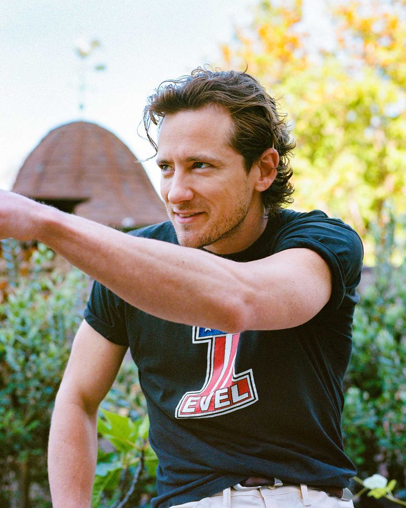 A man in a black T-shirt stands outdoors, pointing. The scene is naturally lit, featuring a barn and greenery in the background.