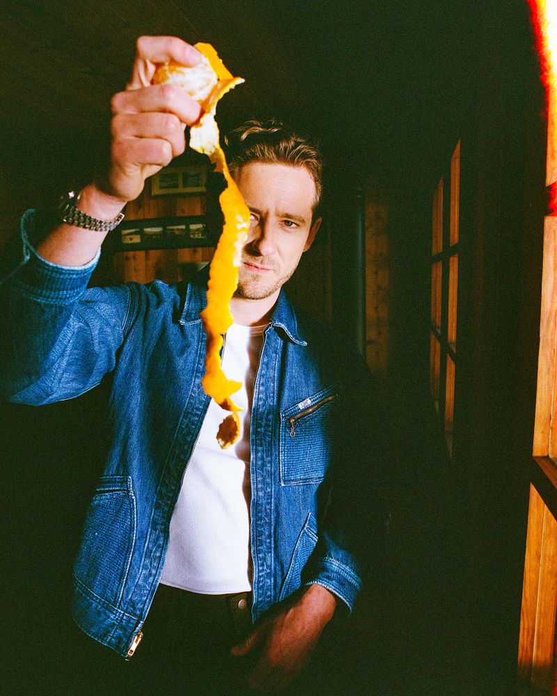 Adult male peeling an orange in an indoor setting, wearing a denim jacket and white t-shirt, with a dark background.