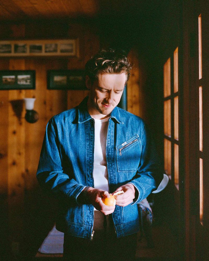Indoor portrait of an adult male in a denim jacket peeling an orange in a room with wood-paneled walls and natural light streaming through a window.